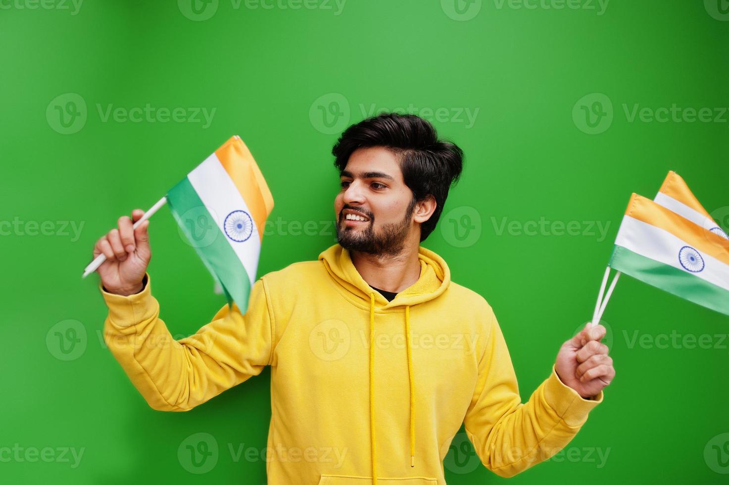 Urban young hipster indian man in a fashionable yellow sweatshirt hold flags of India in hands. Cool south asian guy wear hoodie isolated on green background. photo