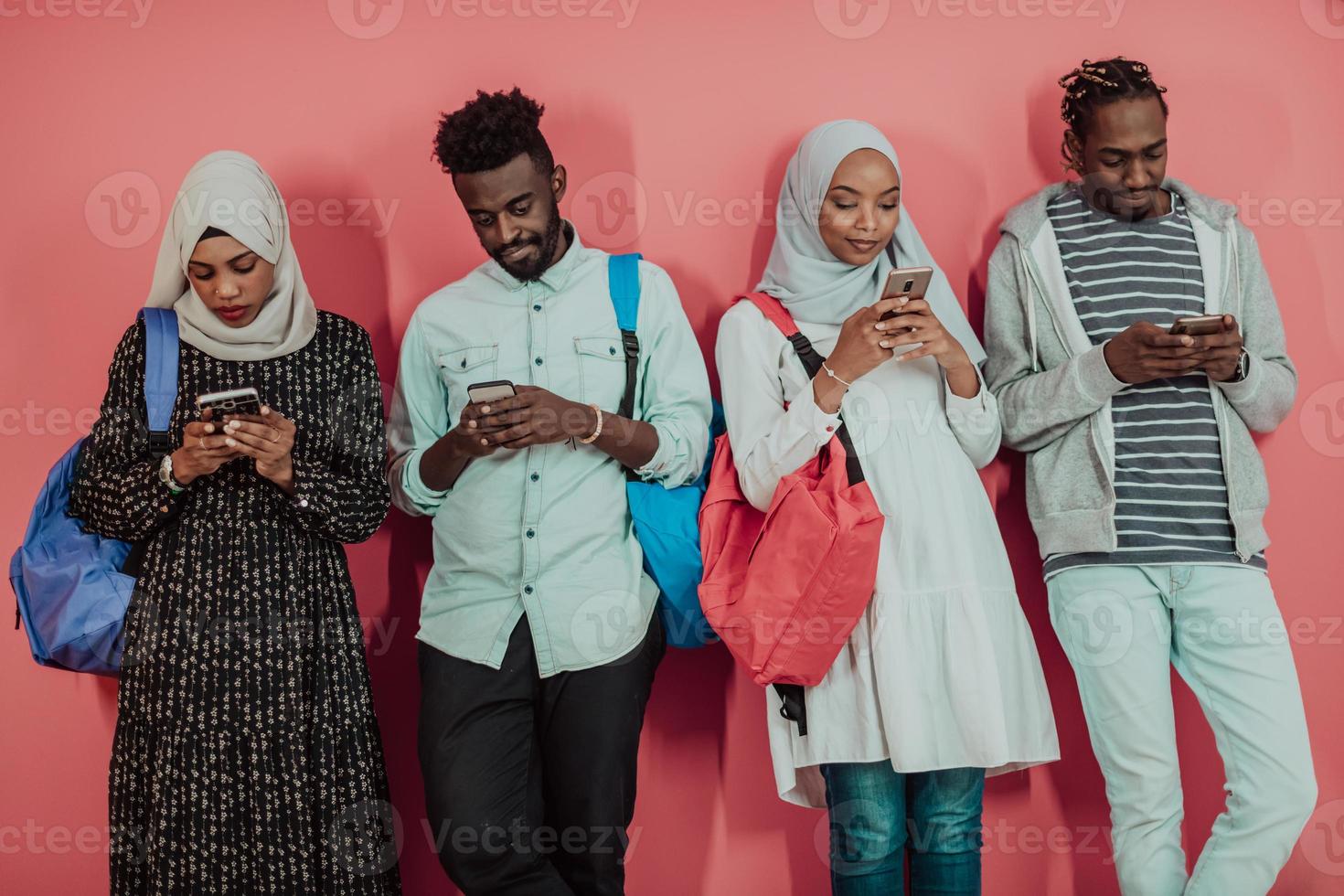 A group of African Muslim students use smartphones while standing in front of a pink background photo