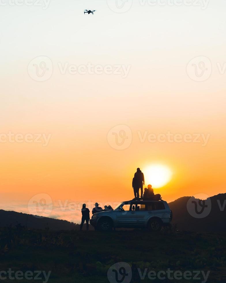 Group of travelers content creators stand by 4wd vehicle together outdoors in nature adventure watch sunset over horizon over cloudscape in wilderness. Exploration and adventure photo