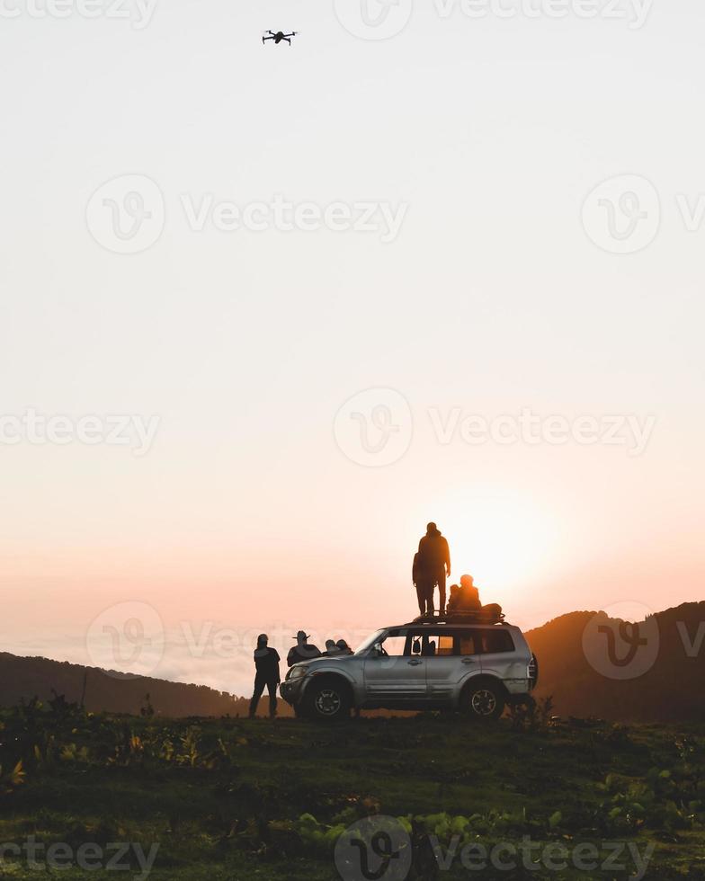 Group of travelers content creators stand by 4wd vehicle together outdoors in nature adventure watch sunset over horizon over cloudscape in wilderness. Exploration and adventure photo