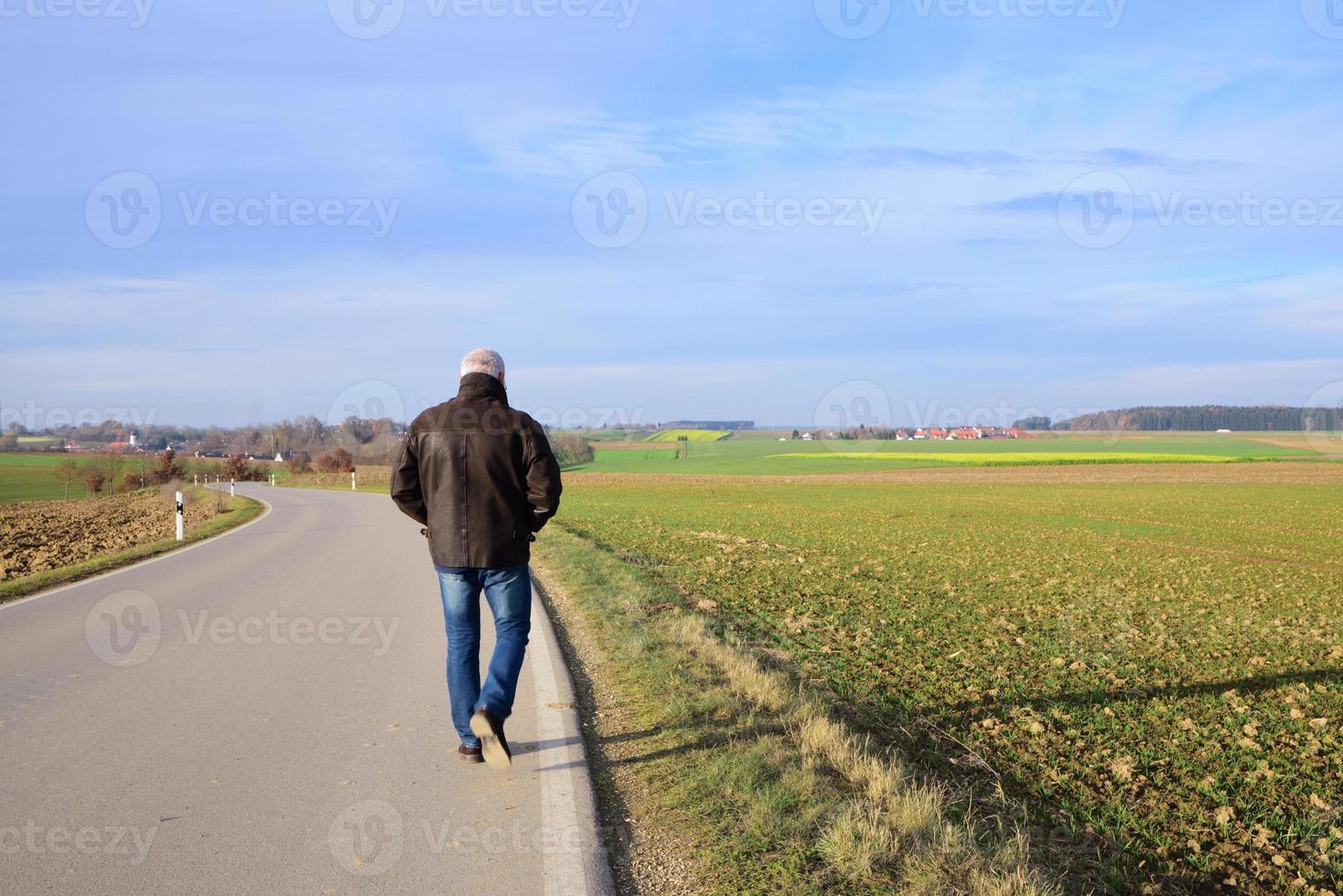 In autumn, a man walks alone on a deserted road that leads over harvested fields, against a blue sky with clouds photo