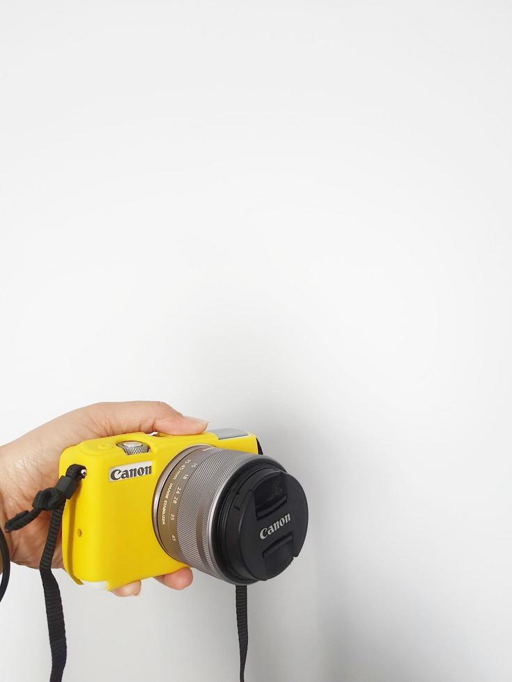 A hand is holding a canon eos m10 camera, this camera is given a yellow rubber protector. photo