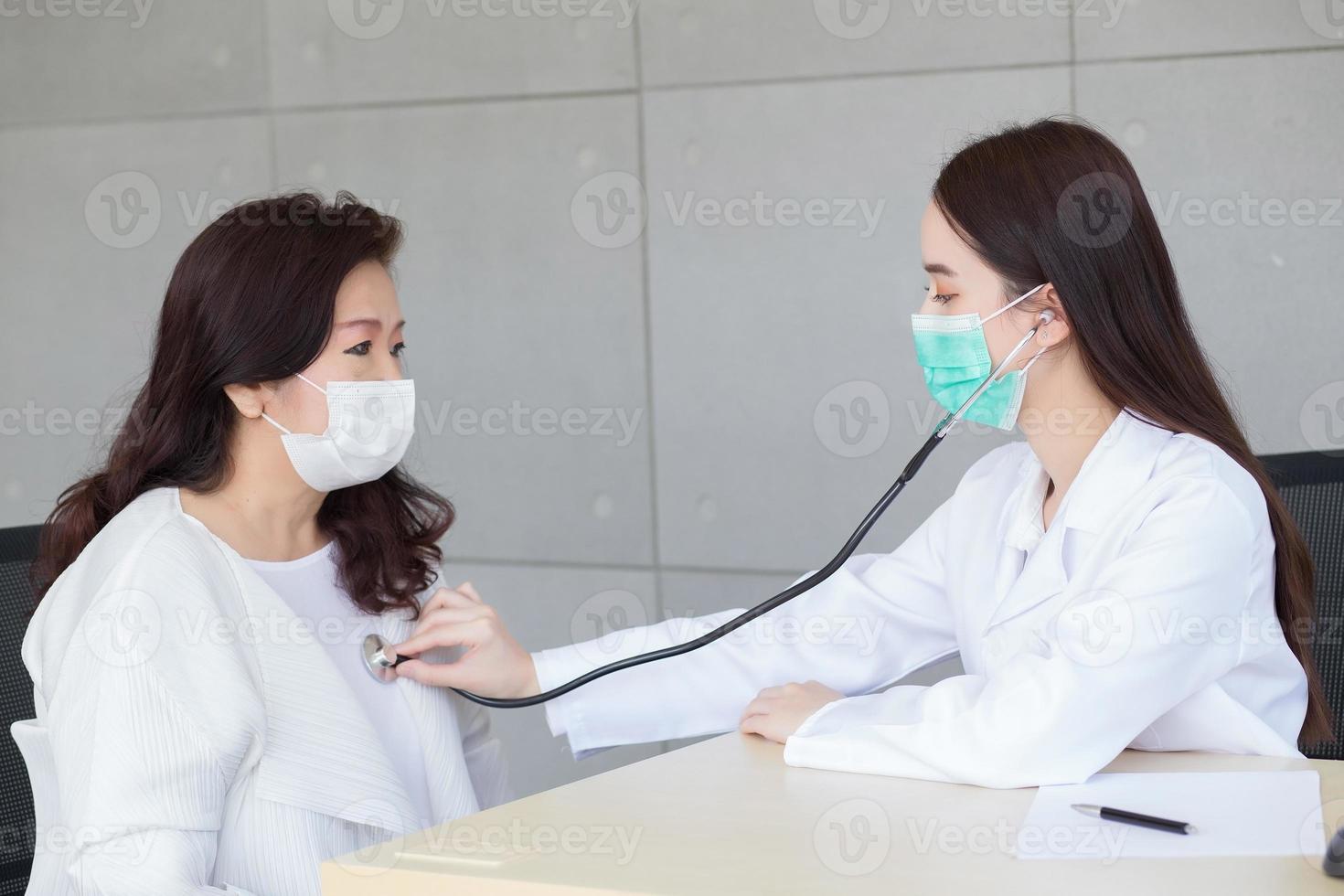 Asian beautiful woman doctor uses stethoscope to check health of lungs and heart or diagnosis symptom of woman  elderly patient while both wear face mask in hospital. photo