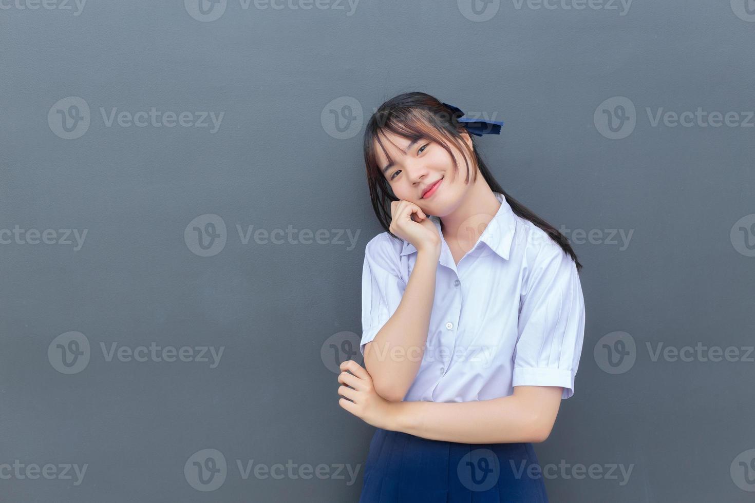 linda estudiante asiática de secundaria con uniforme escolar con sonrisas confiadas mientras mira a la cámara felizmente con gris en el fondo. foto