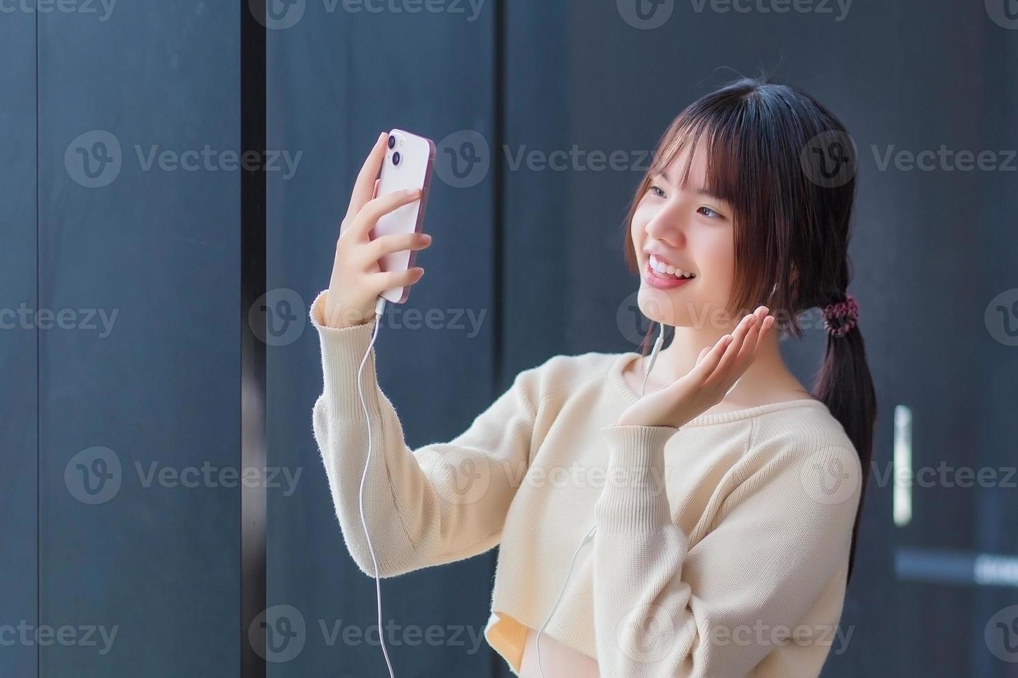 una linda estudiante asiática adolescente que usa una camisa de manga larga está usando su teléfono inteligente y auriculares para hacer una videollamada divertida con sus amigos mientras espera para entrar al salón de clases en la escuela. foto