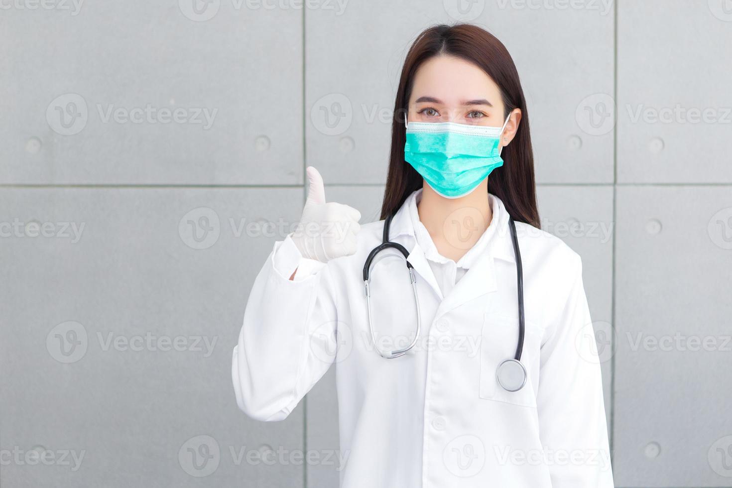 Asian woman doctor who wears medical coat and face mask shows thump up as good sign in health protection concept at hospital. photo