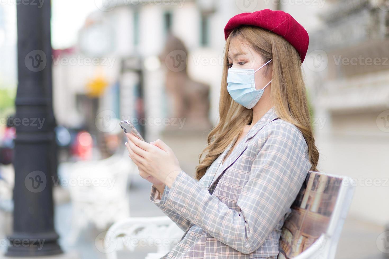 Asian beautiful girl wearing suit and red cap holds smartphone in her hands while sits on bench in park while wearing medical face mask in new normal lifestyle concept. photo