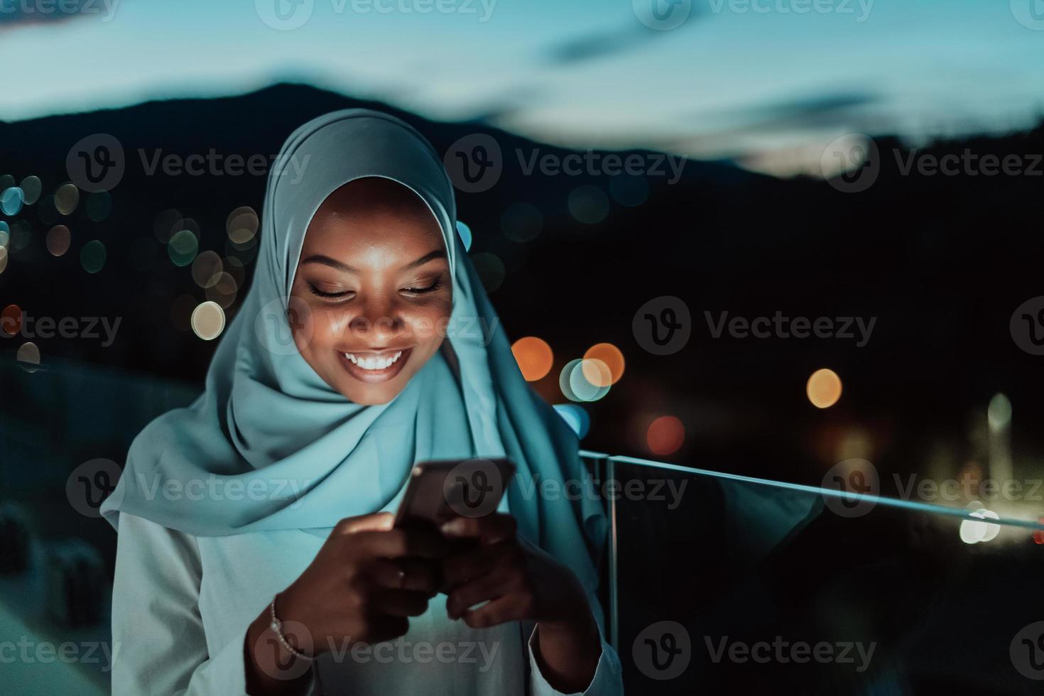 joven mujer musulmana con velo de bufanda en la calle urbana de la ciudad por la noche enviando mensajes de texto en un teléfono inteligente con la luz de la ciudad en el fondo. foto