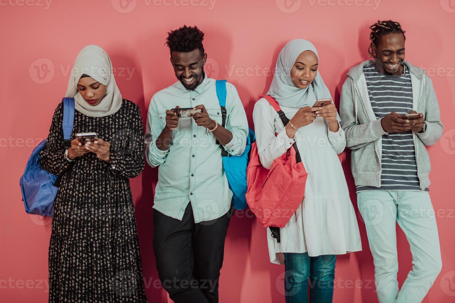 A group of African Muslim students use smartphones while standing in front of a pink background photo