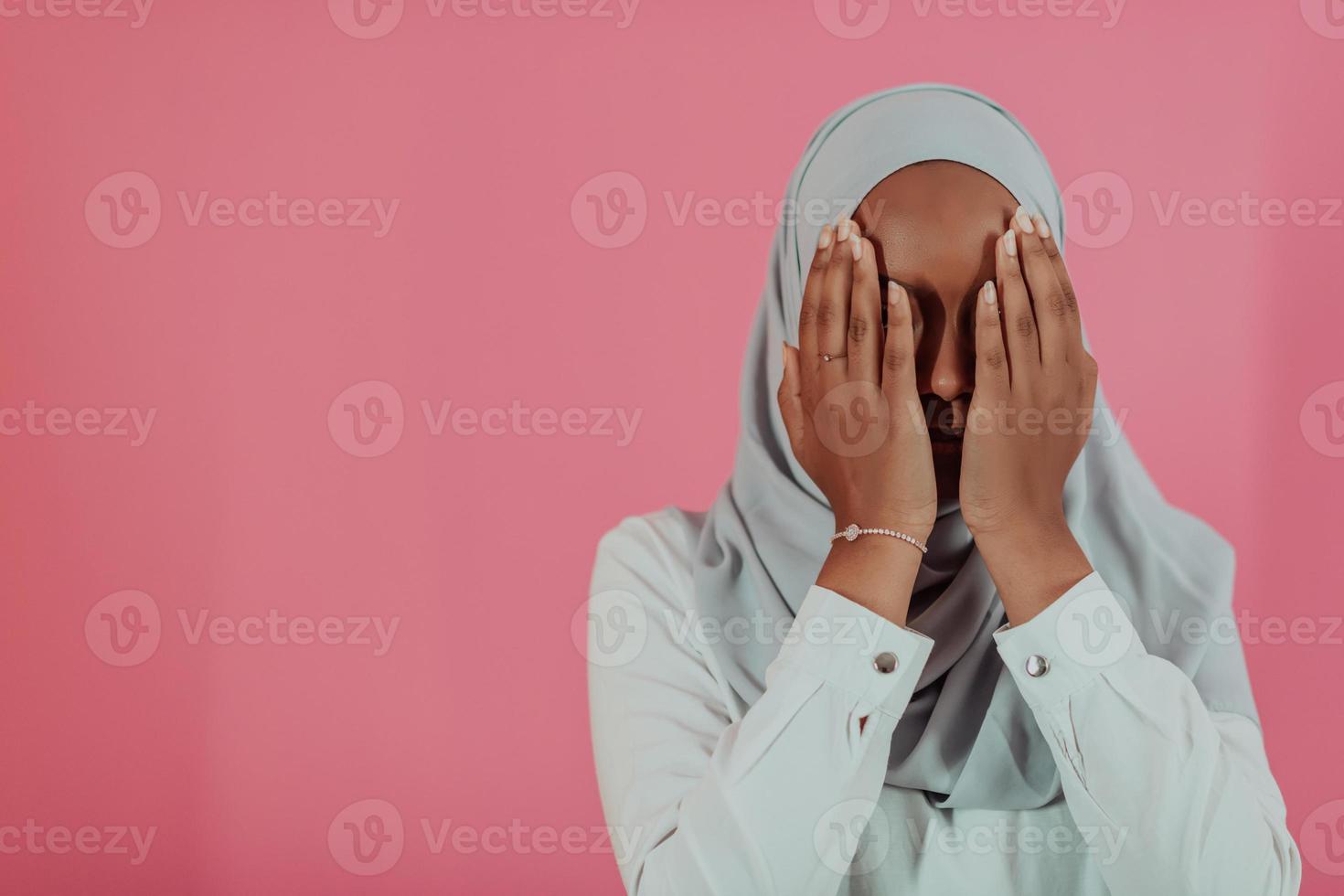 Modern African Muslim woman makes traditional prayer to God, keeps hands in praying gesture, wears traditional white clothes, has serious facial expression, isolated over plastic pink background photo