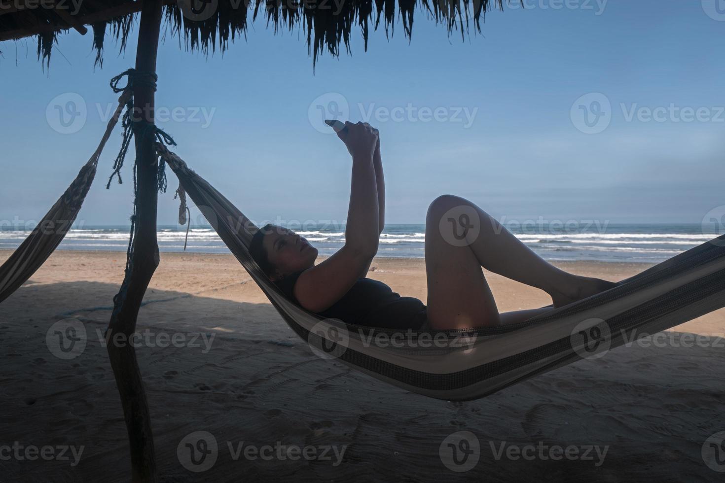 bella joven hispana vestida con una camiseta azul descansando en una hamaca tomando selfies con su smartphone en la playa foto