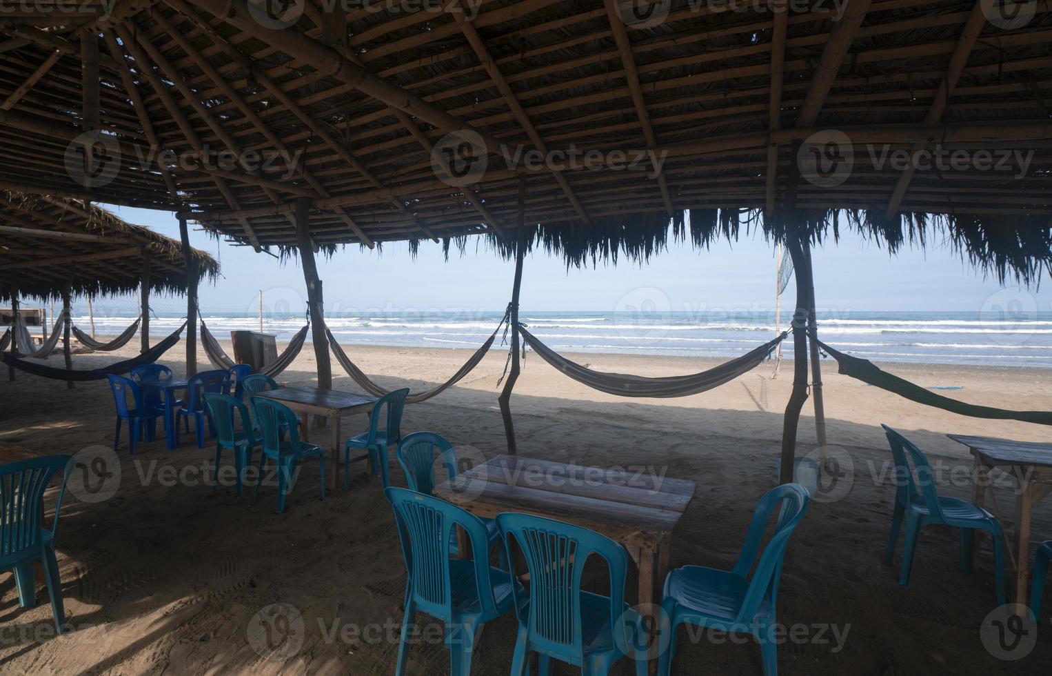 View of a rustic restaurant without people, with a bamboo roof, wooden tables, plastic chairs and hammocks hanging from poles by the beach photo