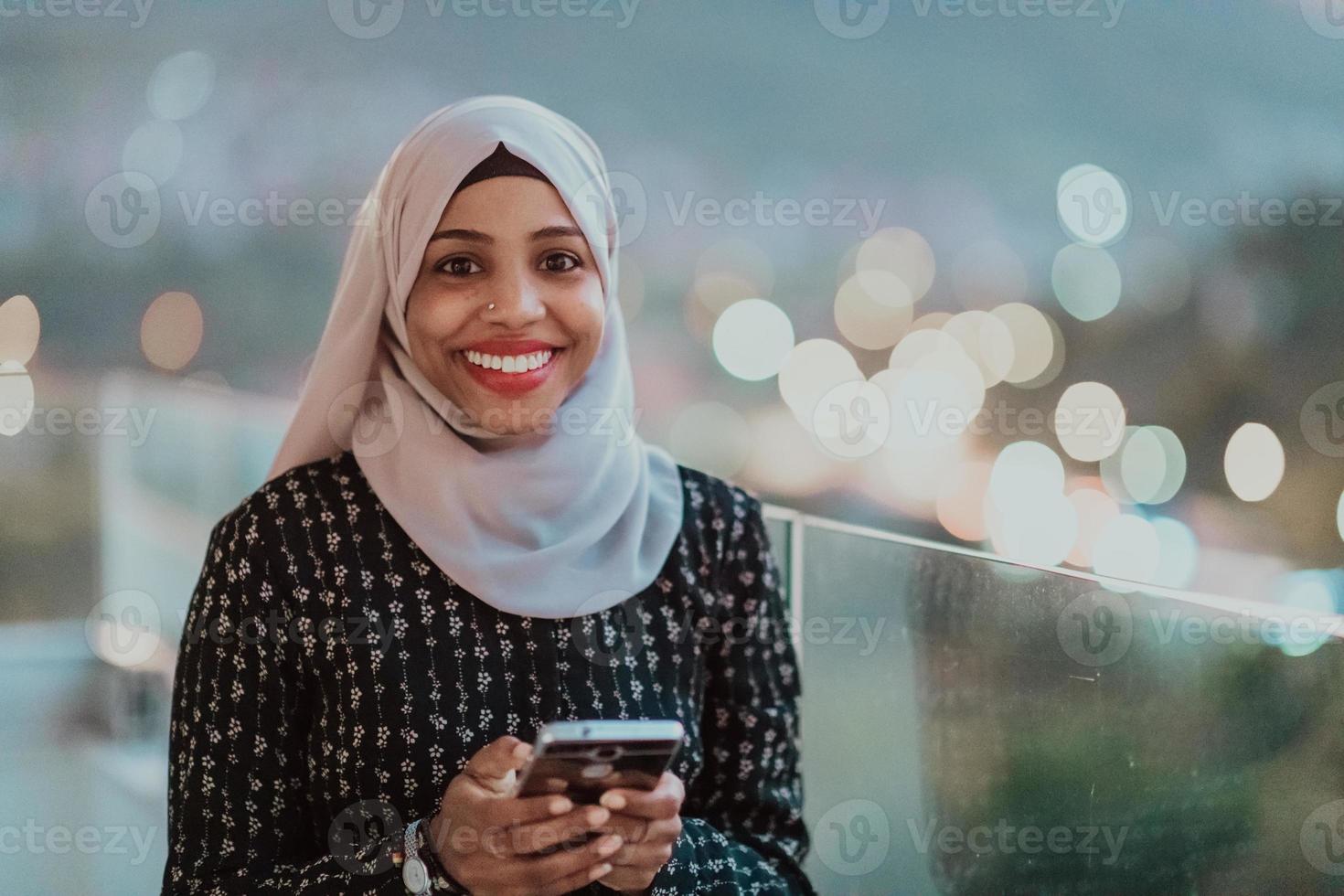 joven mujer musulmana con velo de bufanda en la calle urbana de la ciudad por la noche enviando mensajes de texto en un teléfono inteligente con la luz de la ciudad en el fondo. foto