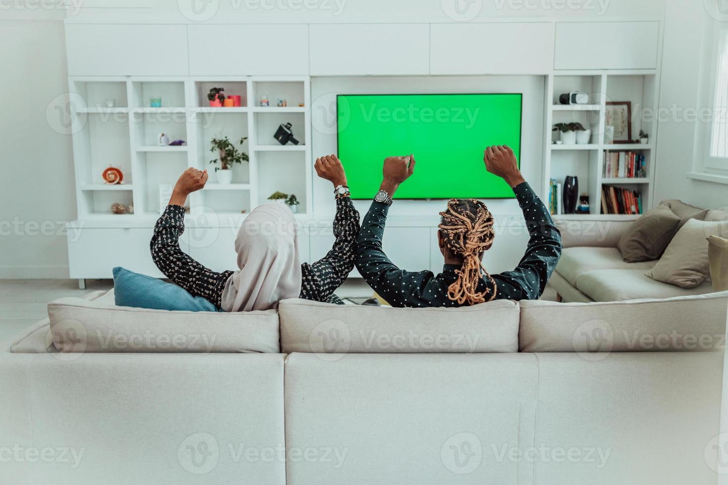 African Couple Sitting On Sofa Watching TV Together Chroma Green Screen Woman Wearing Islamic Hijab Clothes photo