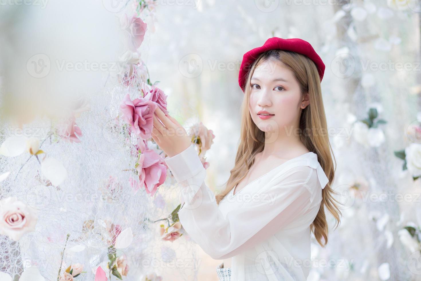 hermosa mujer asiática con el pelo largo de bronce en camisa blanca de manga larga sonríe y se para en el jardín de flores de rosas blancas como un tema natural y lujoso foto