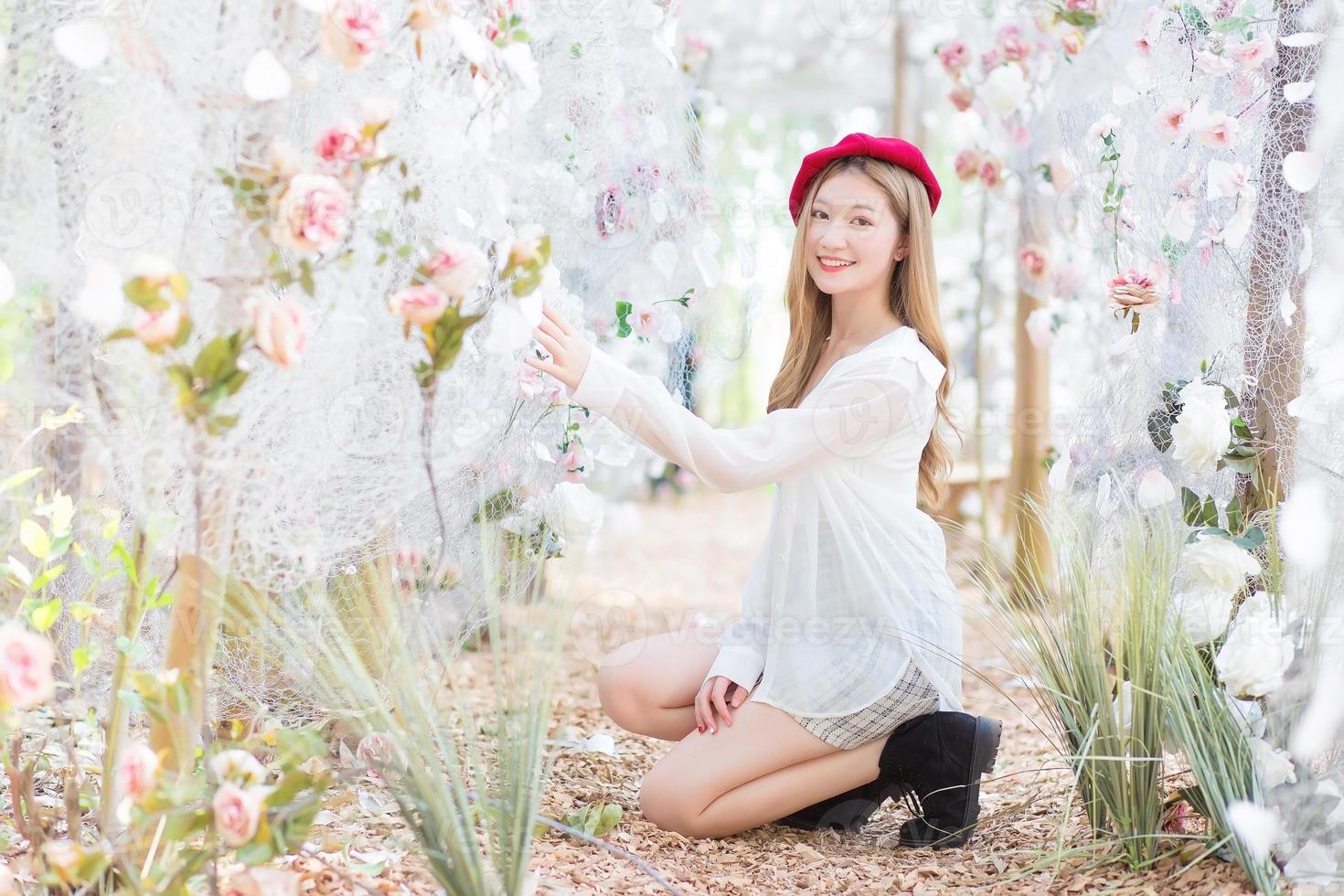 Asian woman has long bronze hair and wears red cap, white dress and black boots while she sits among flower garden in winter season. photo