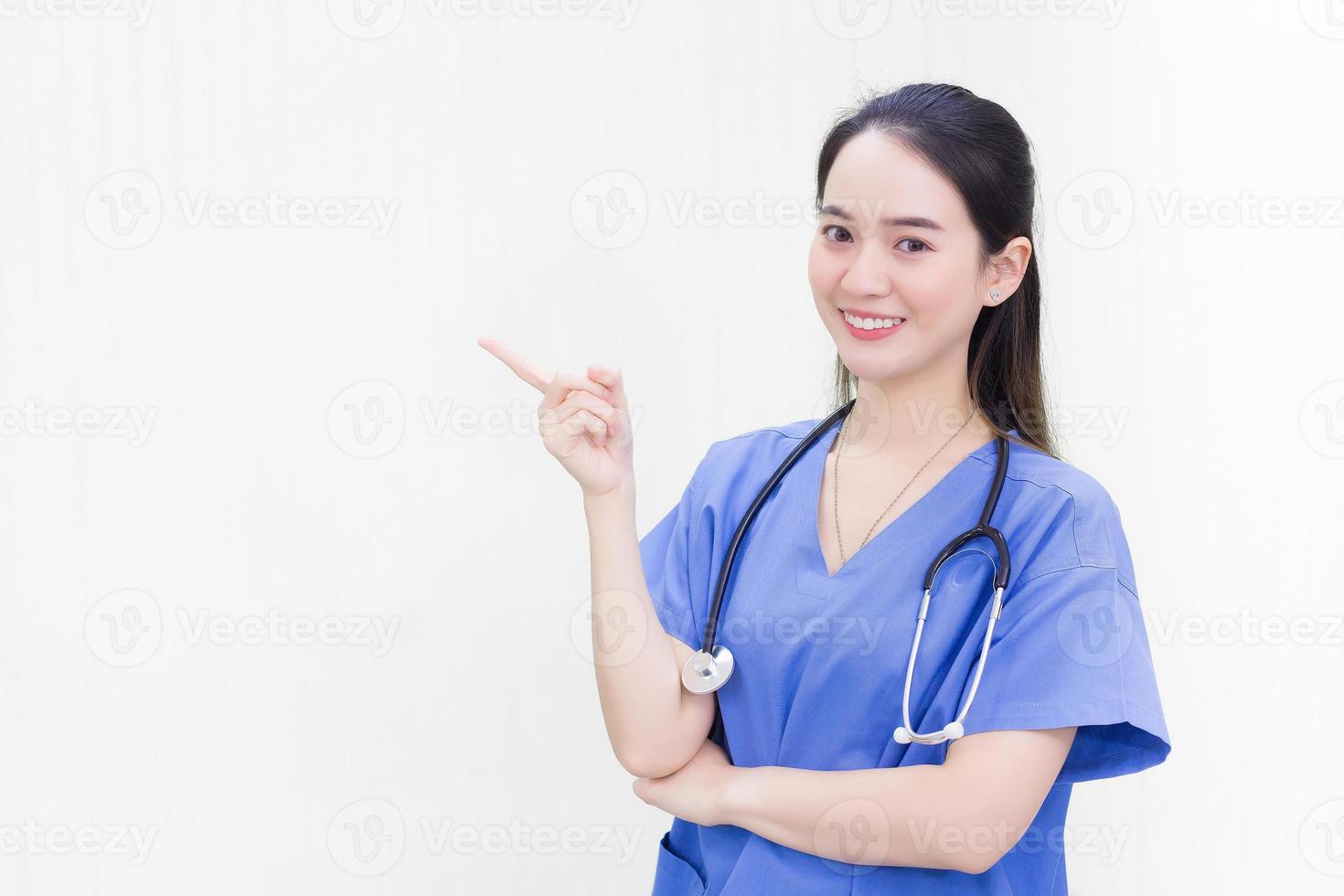 una hermosa doctora asiática con uniforme azul se pone de pie y sonríe mientras señala la parte superior sobre un fondo blanco. foto