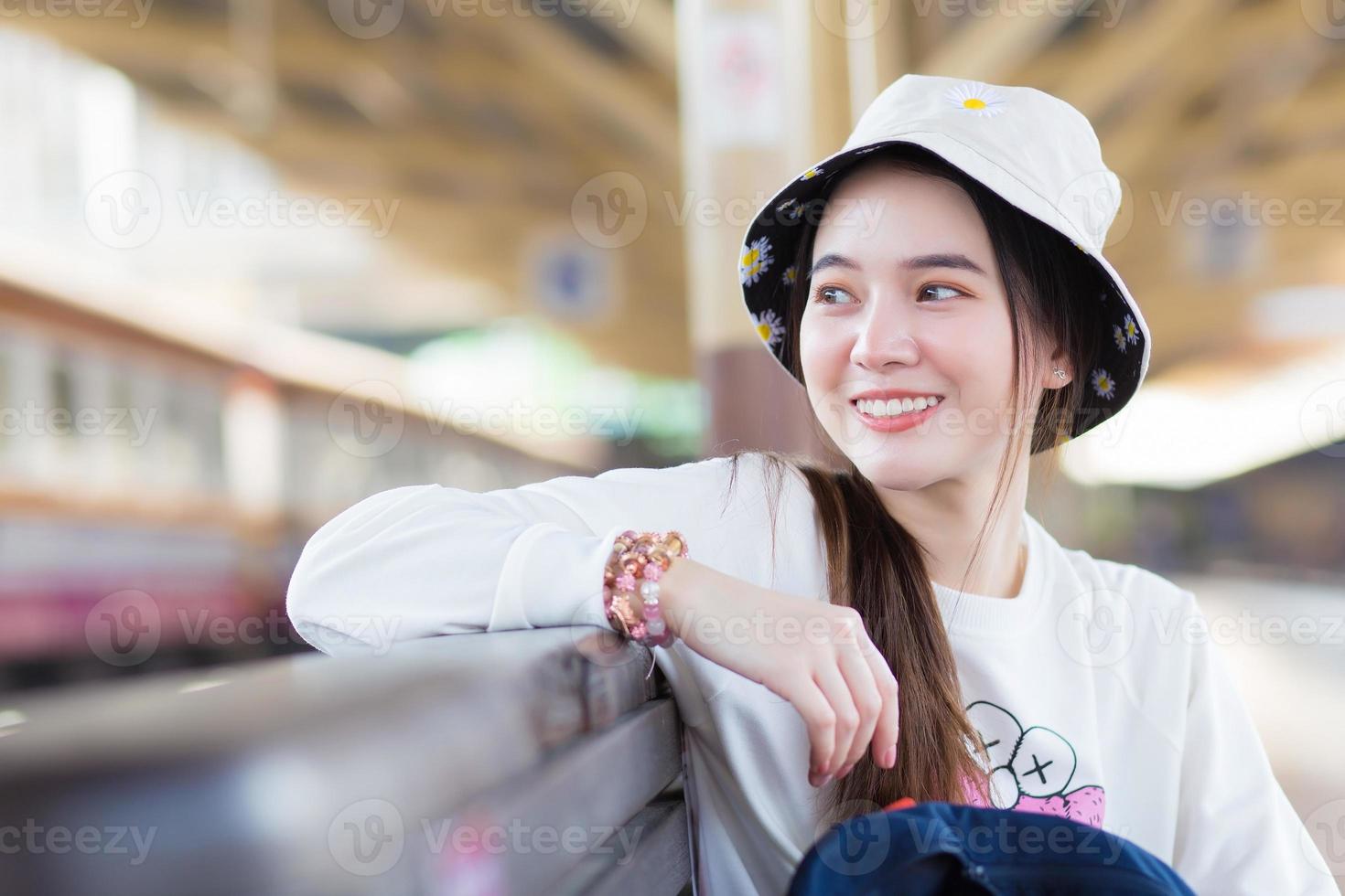 Asian beautiful girl in a long-sleeved white shirt and a hat sits happy smilie in the train station waiting for the train to arrive. photo