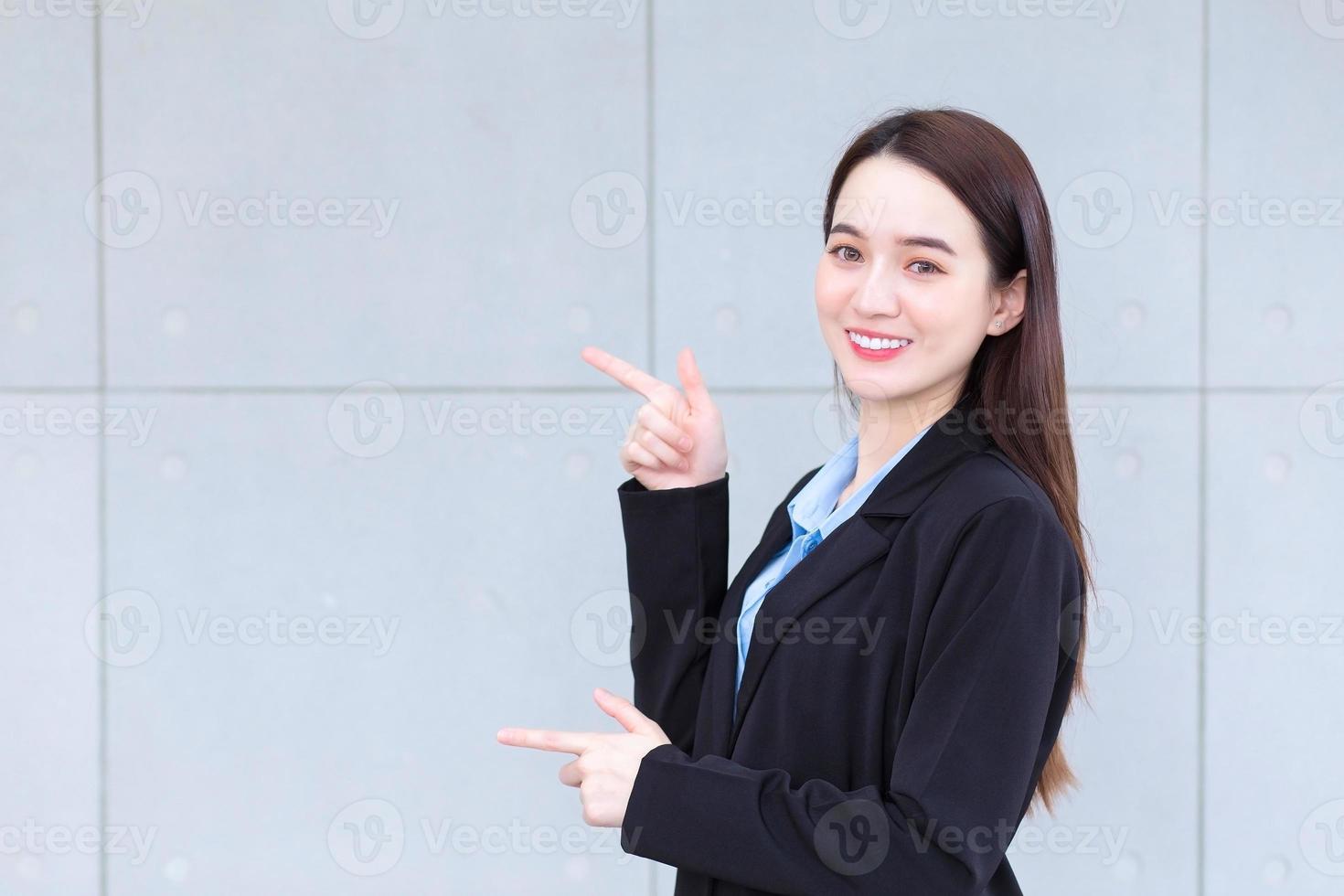una trabajadora asiática que viste traje negro señala con la mano para presentar algo en la pared. foto