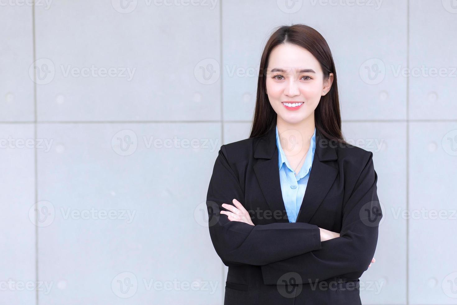 mujer trabajadora asiática que tiene el pelo largo viste traje negro formal con camisa azul mientras cruza el brazo y sonríe alegremente. foto