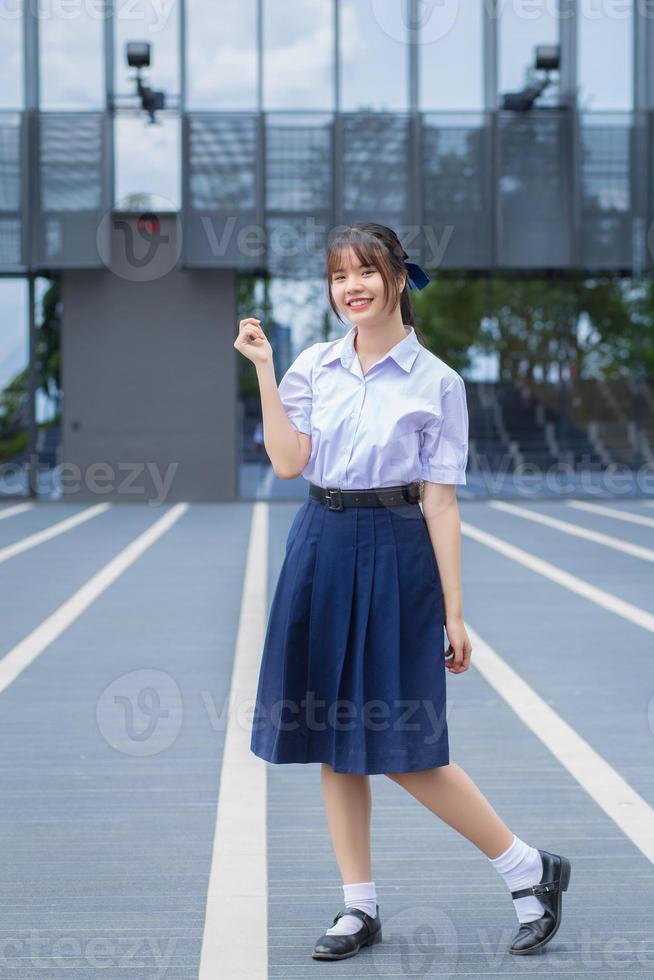 una linda estudiante asiática de secundaria con el uniforme escolar se pone de pie y sonríe alegremente mientras confía en el edificio gris como fondo. foto