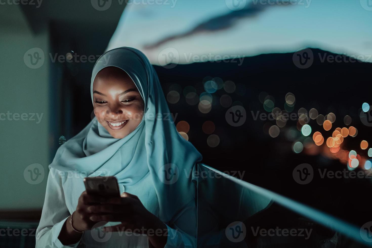 joven mujer musulmana con velo de bufanda en la calle urbana de la ciudad por la noche enviando mensajes de texto en un teléfono inteligente con la luz de la ciudad en el fondo. foto