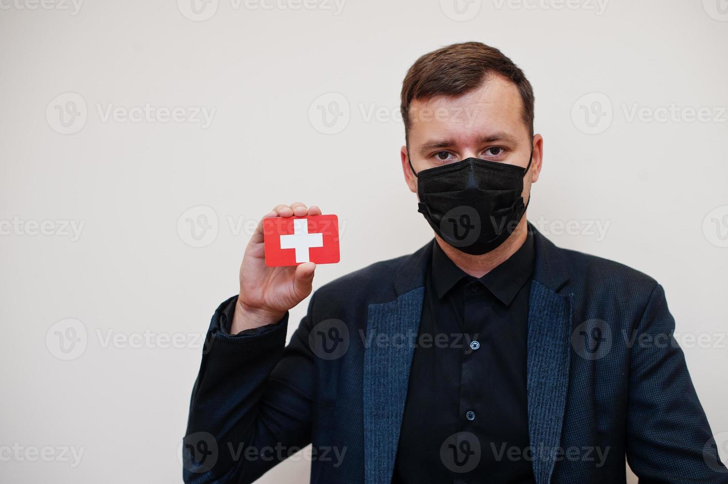 European man wear black formal and protect face mask, hold Switzerland flag card isolated on white background. Europe coronavirus Covid country concept. photo