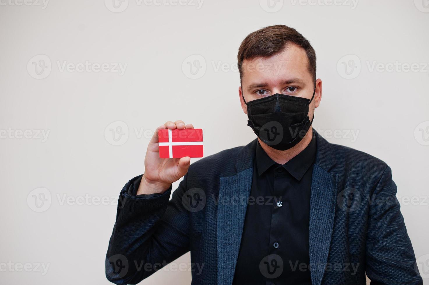 European man wear black formal and protect face mask, hold Denmark flag card isolated on white background. Europe coronavirus Covid country concept. photo