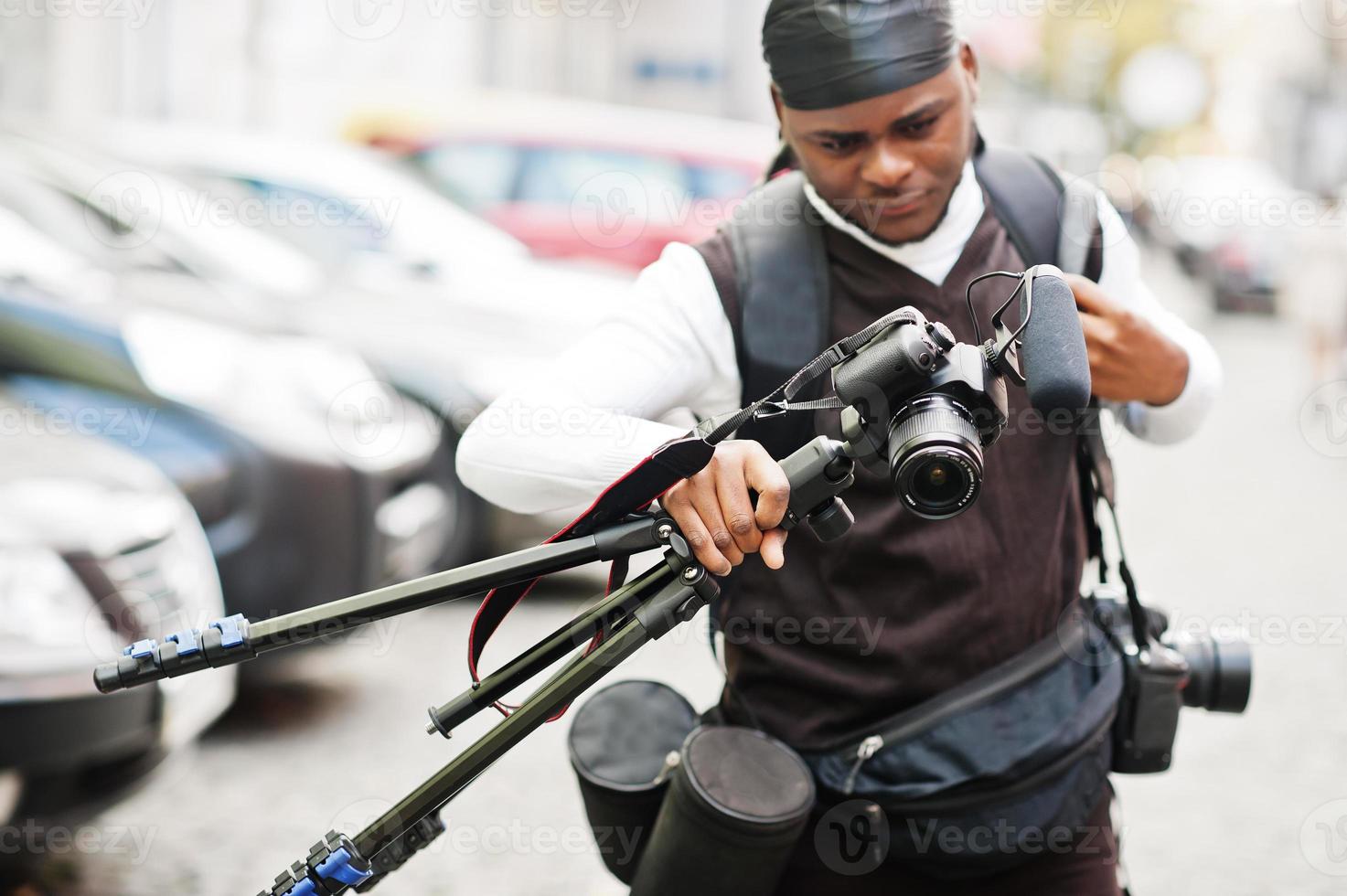 Young professional african american videographer holding professional camera with tripod pro equipment. Afro cameraman wearing black duraq making a videos. photo