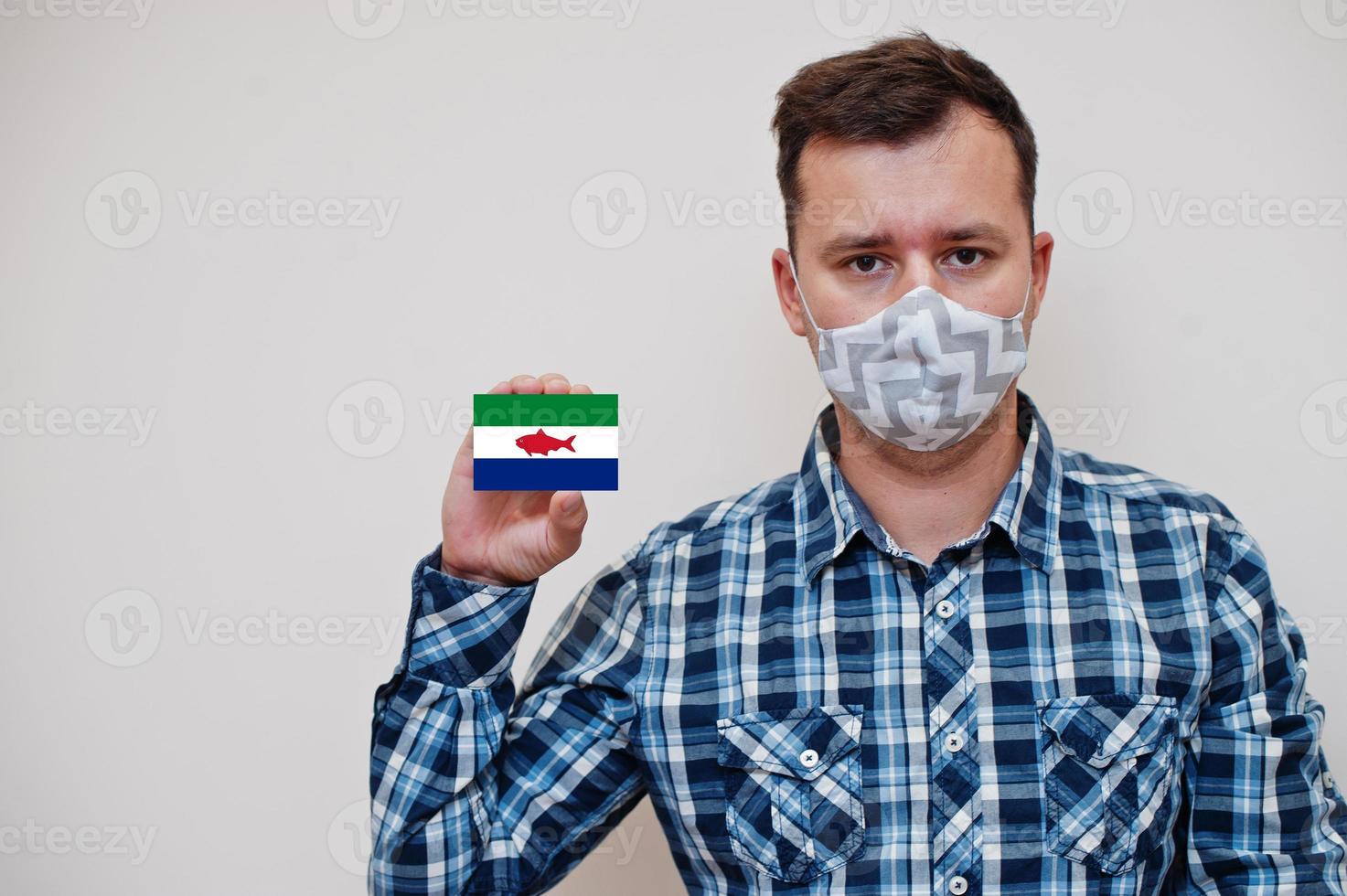 Man in checkered shirt show Federal Dependencies of Venezuela flag card in hand, wear protect mask isolated on white background. American countries Coronavirus concept. photo