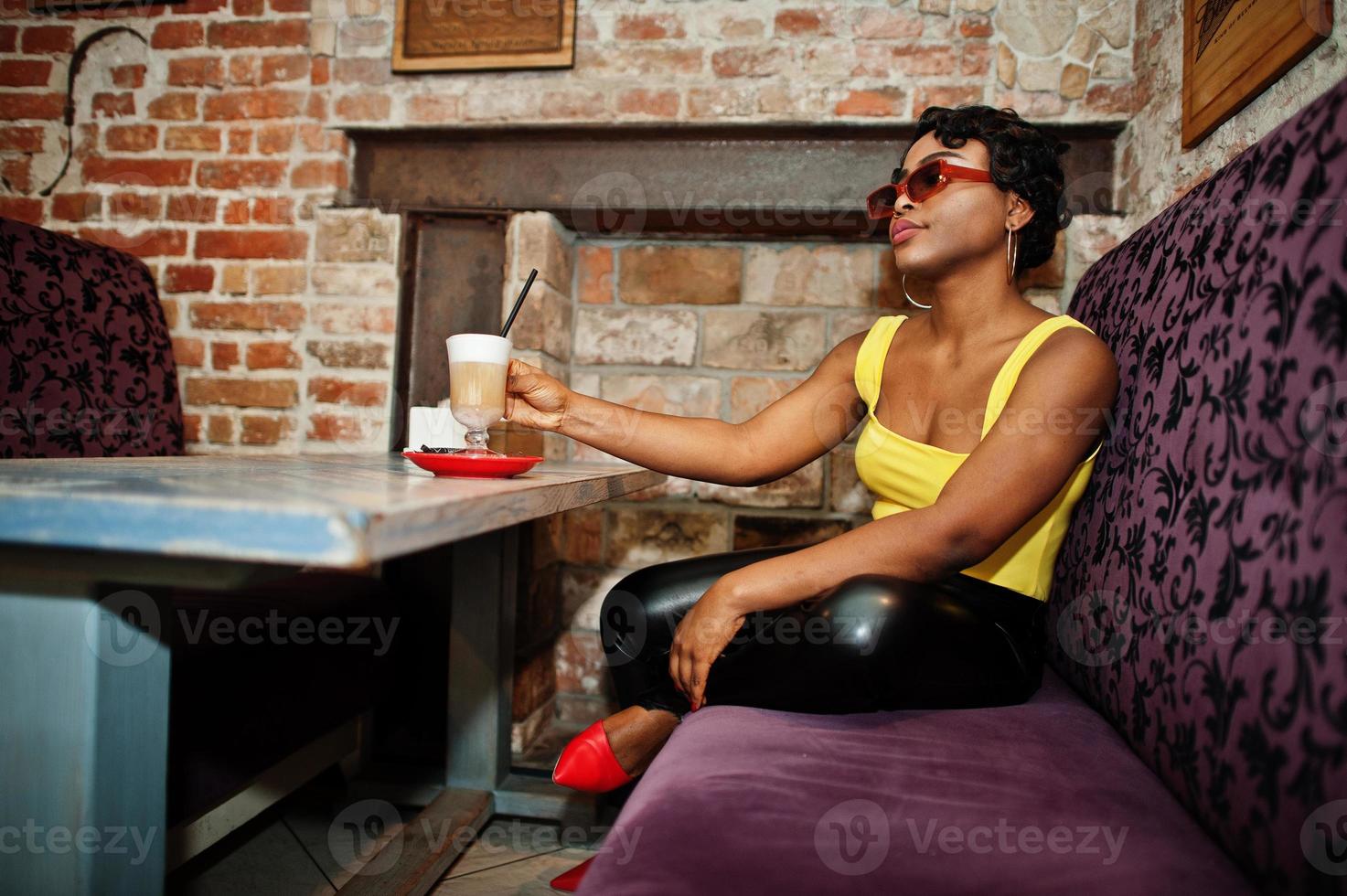 Stunning African American women in yellow top and black leather pants pose at pub with coffee. photo