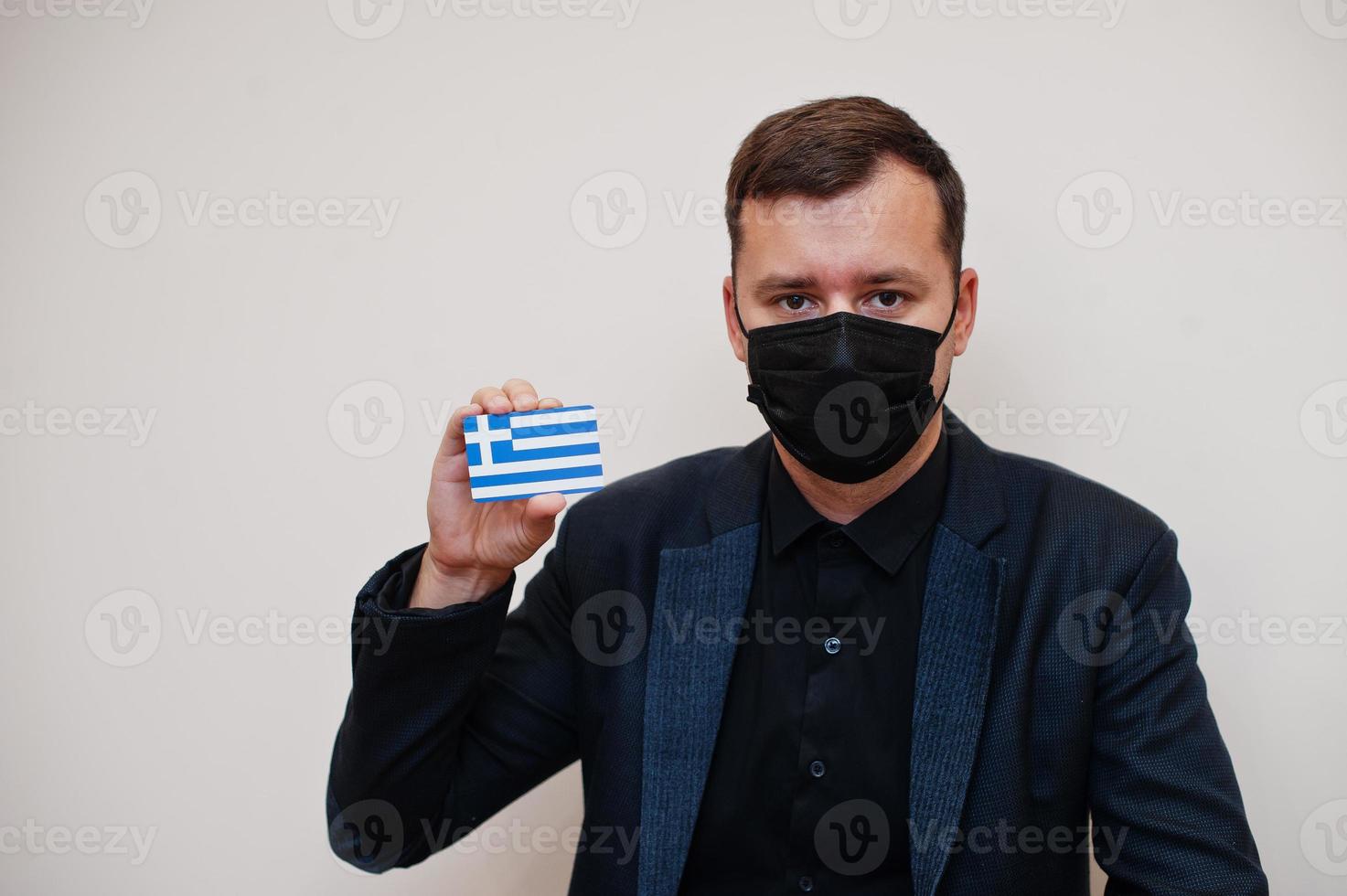 European man wear black formal and protect face mask, hold Greece flag card isolated on white background. Europe coronavirus Covid country concept. photo