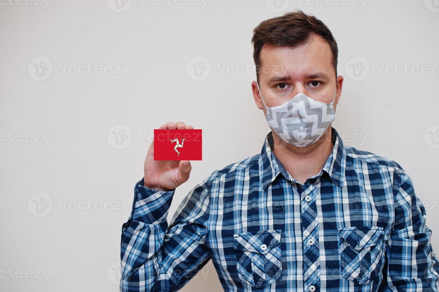 Man in checkered shirt show Isle of Man flag card in hand, wear protect mask isolated on white background. Europe countries Coronavirus concept. photo