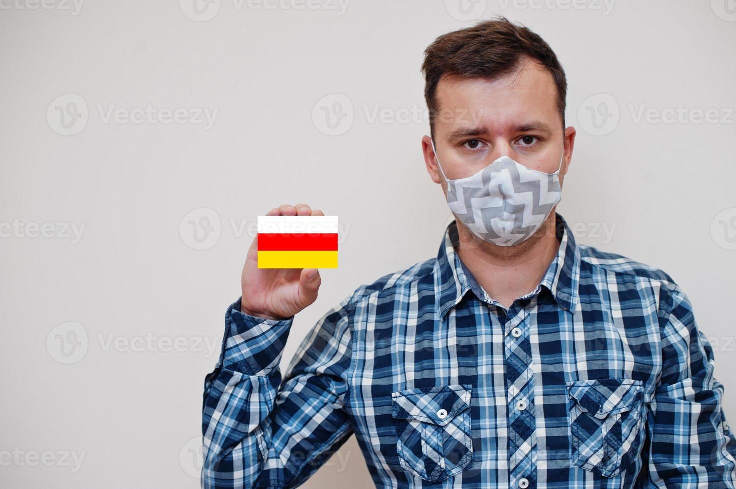 Man in checkered shirt show South Ossetia flag card in hand, wear protect mask isolated on white background. Europe countries Coronavirus concept. photo