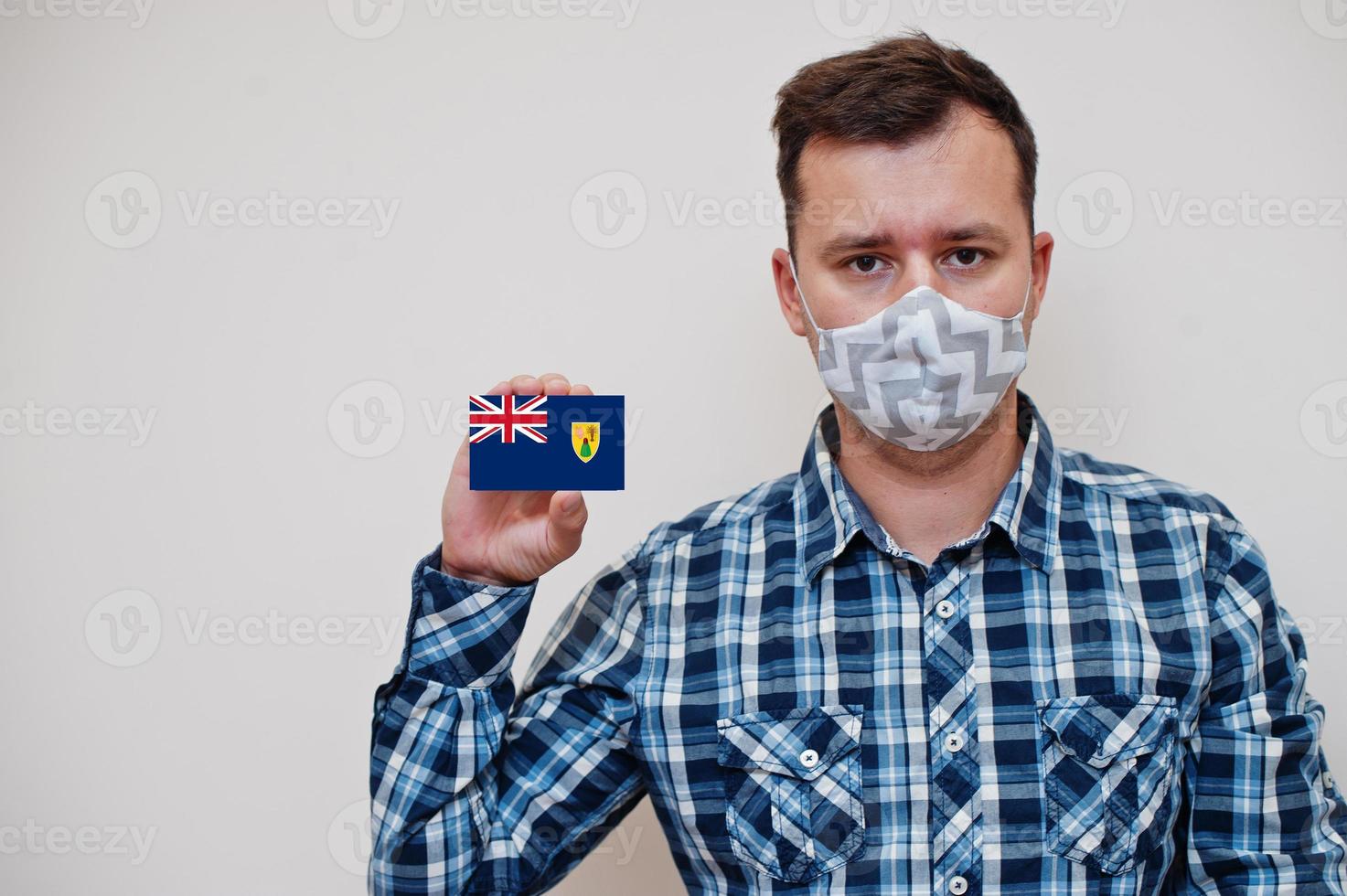 Man in checkered shirt show Turks and Caicos Islands flag card in hand, wear protect mask isolated on white background. American countries Coronavirus concept. photo