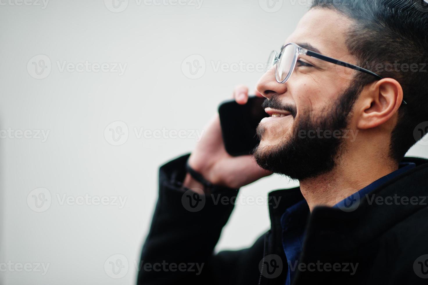 Middle eastern entrepreneur wear black coat and blue shirt, eyeglasses standing against white wall, smoking cigarette and speak by phone. photo