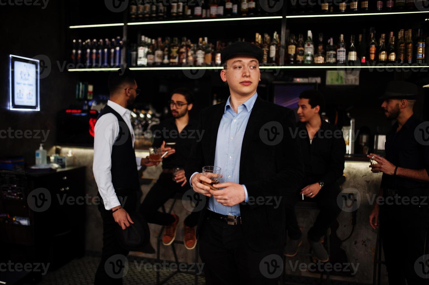 hombre árabe con estilo contra un grupo de gánsteres guapos retro bien vestidos pasan tiempo en el club, bebiendo en el mostrador del bar. despedida de soltero multiétnico de la mafia en el restaurante. foto