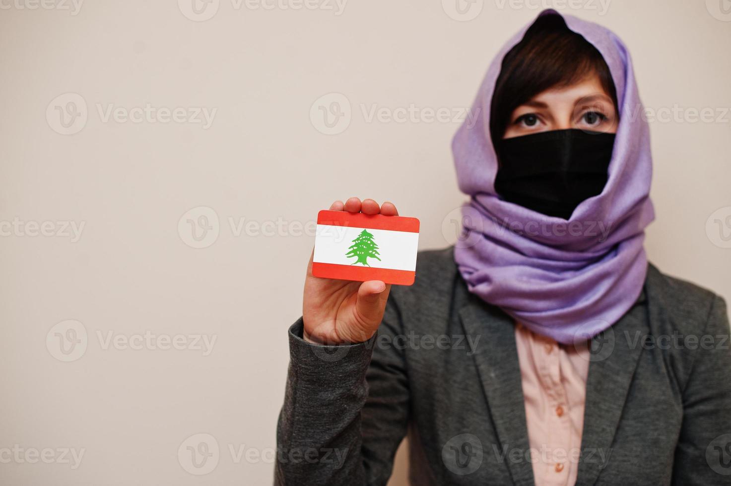 Portrait of young muslim woman wearing formal wear, protect face mask and hijab head scarf, hold Lebanon flag card against isolated background. Coronavirus country concept. photo