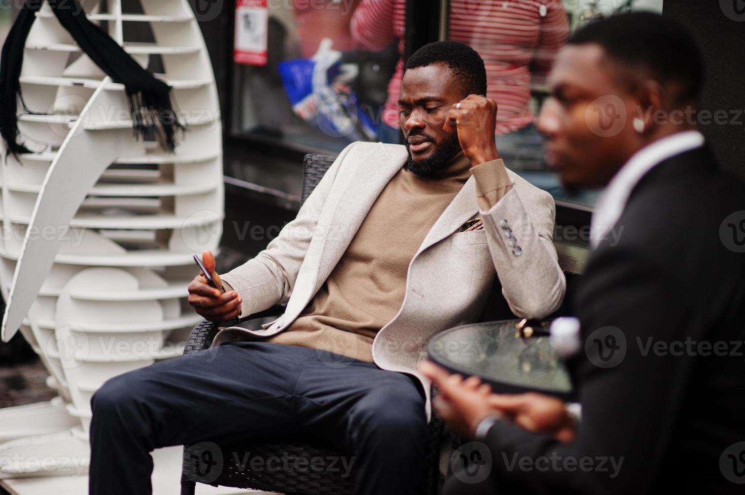 Two fashion black men sitting outdoor with mobile phone. Fashionable portrait of african american male models. Wear suit, coat and hat. photo