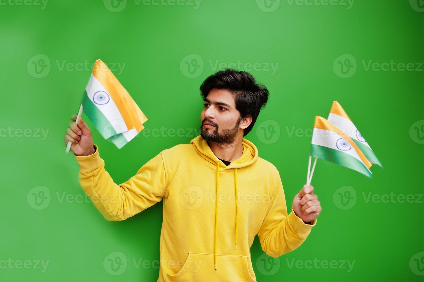 Urban young hipster indian man in a fashionable yellow sweatshirt hold flags of India in hands. Cool south asian guy wear hoodie isolated on green background. photo