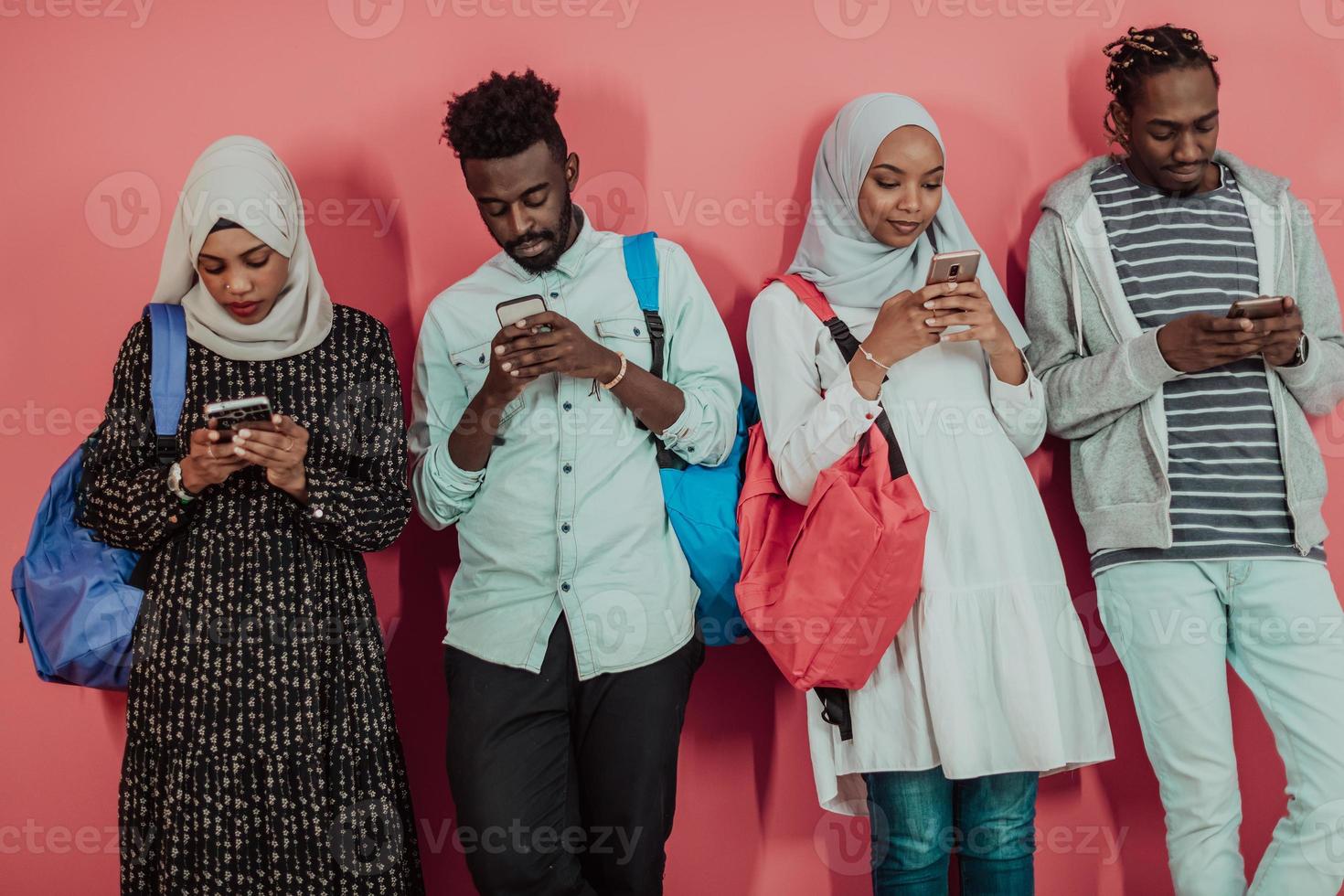 A group of African Muslim students use smartphones while standing in front of a pink background photo