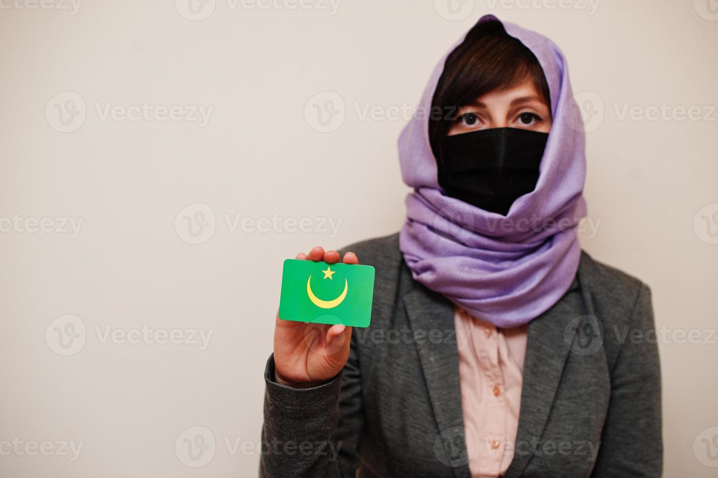 Portrait of young muslim woman wearing formal wear, protect face mask and hijab head scarf, hold Mauritania flag card against isolated background. Coronavirus country concept. photo