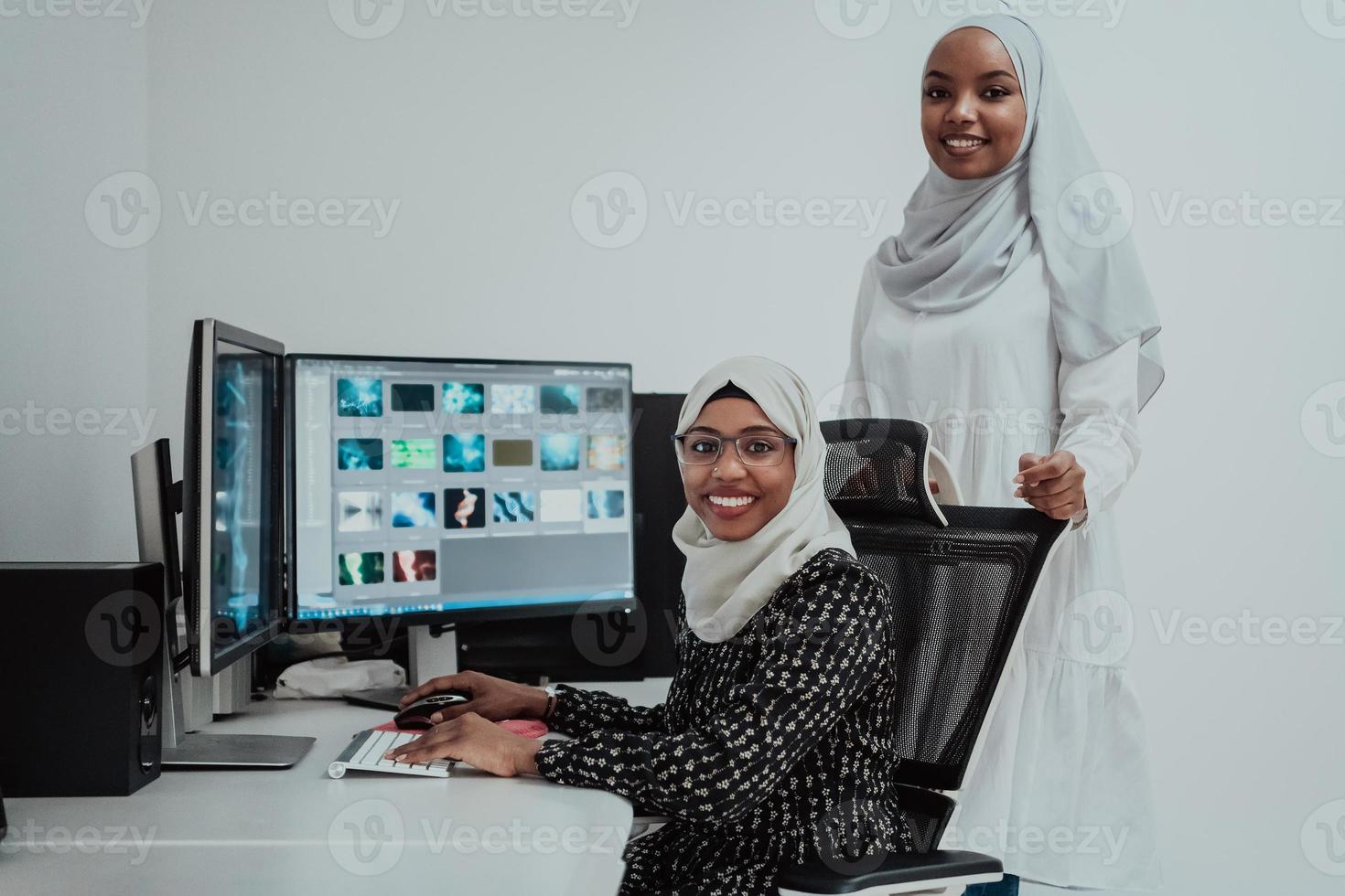 Friends at the office two young Afro American modern Muslim businesswomen wearing scarf in creative bright office workplace with a big screen photo