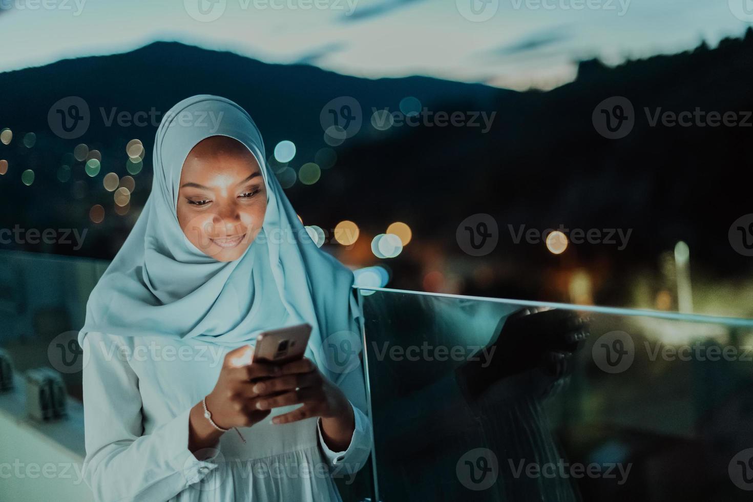 Young Muslim woman wearing scarf veil on urban city street at night texting on a smartphone with bokeh city light in the background. photo