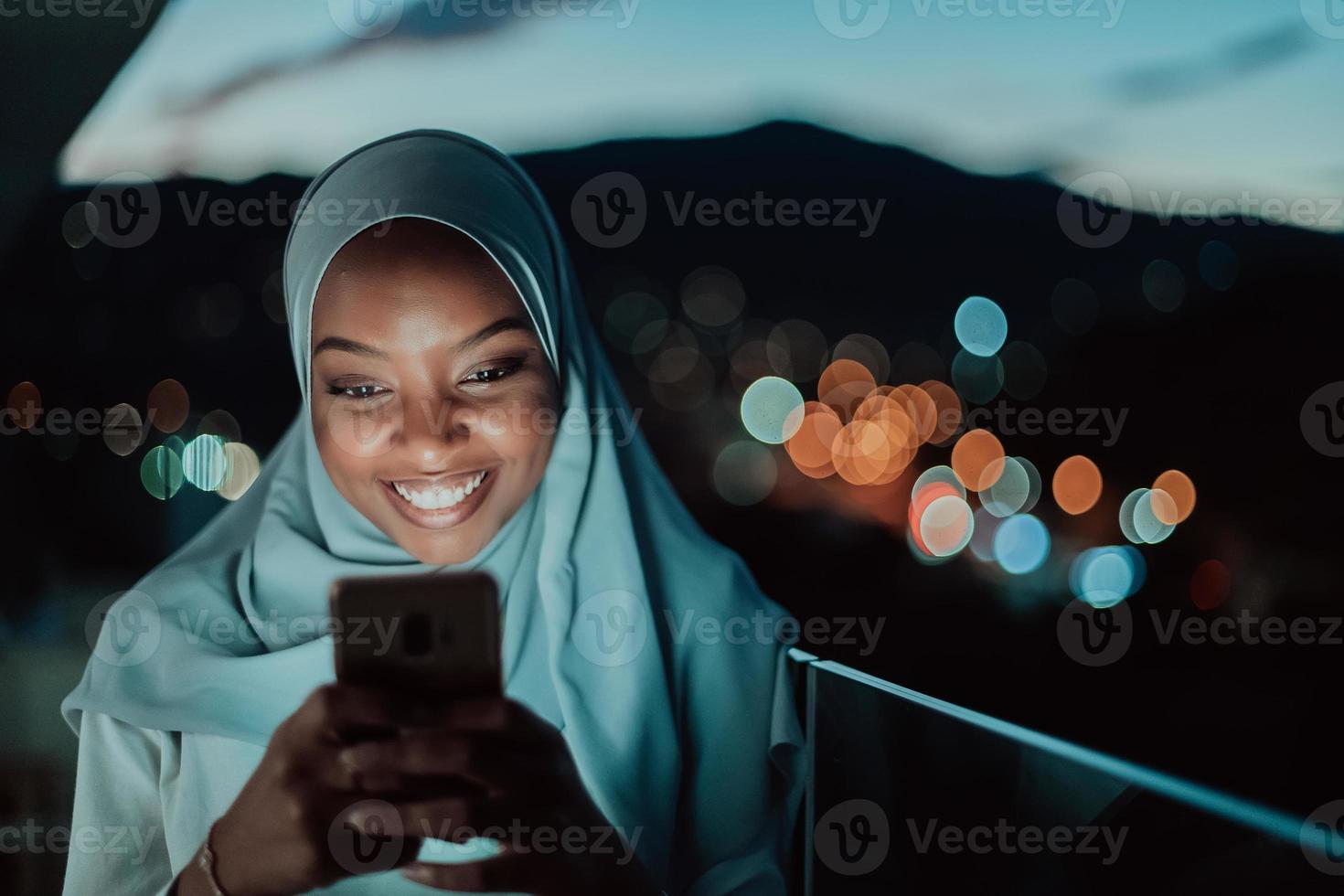 joven mujer musulmana con velo de bufanda en la calle urbana de la ciudad por la noche enviando mensajes de texto en un teléfono inteligente con la luz de la ciudad en el fondo. foto