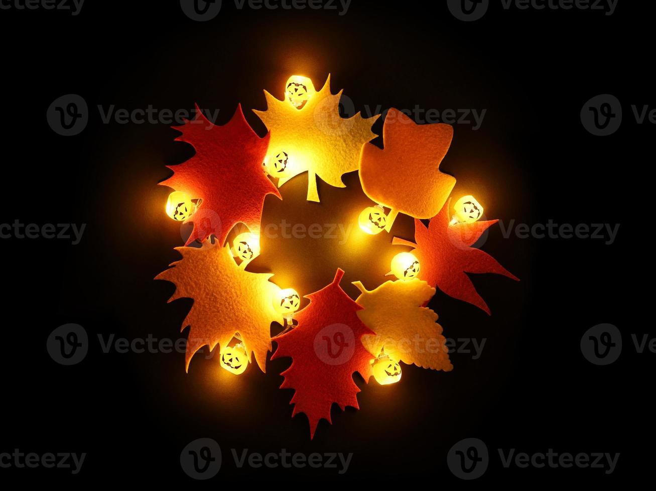 Magic wreath made of colorful tree leaves cut out felt and Halloween garland lights of glowing orange pumpkins on black paper background. Festive holidays Halloween or Thanksgiving decor concept. photo