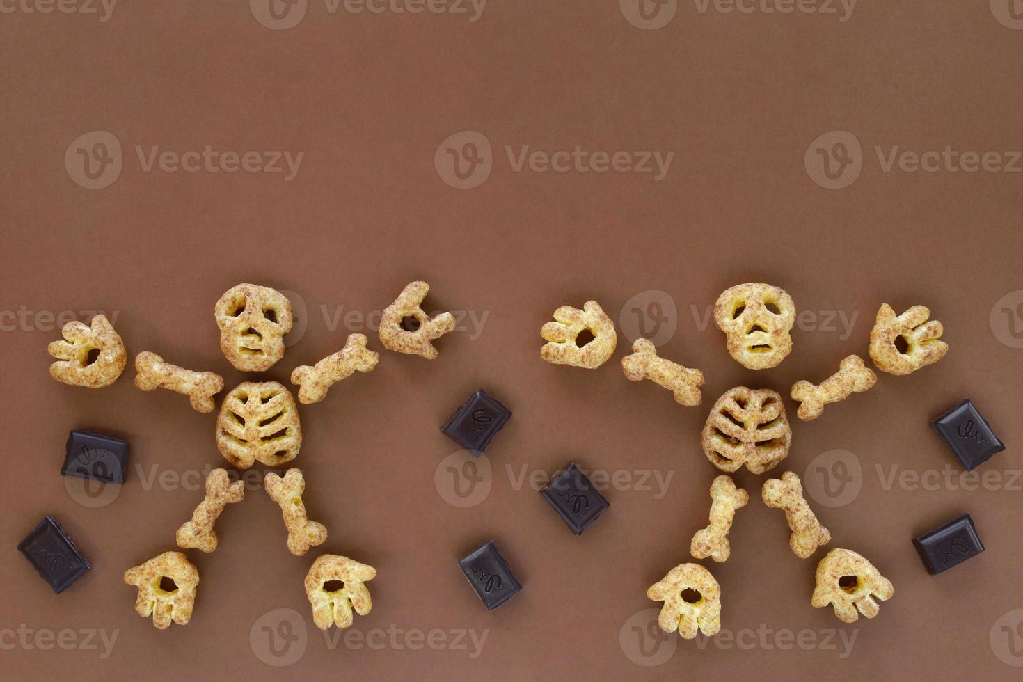 Puffed corn snacks Halloween treat in skeleton form on brown background. Dancing skeleton figures made of parts - skull, rib cage, hands, feet, bones. Pieces of chocolate around. photo