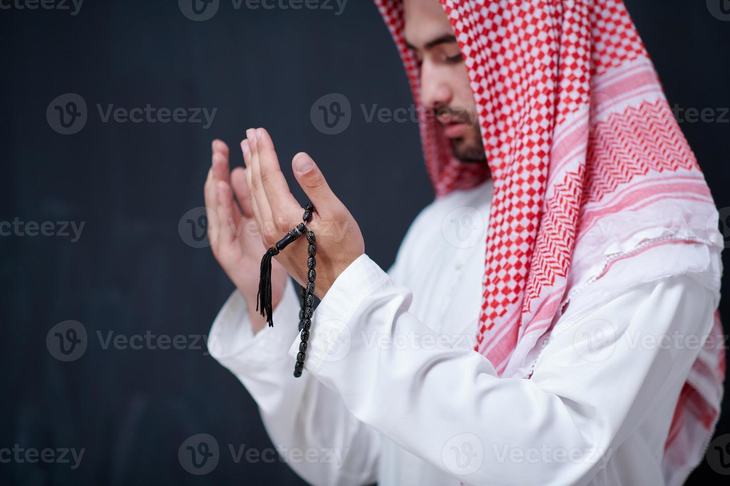 arabian man making traditional prayer to God, keeps hands in praying gesture in front of black chalkboard photo