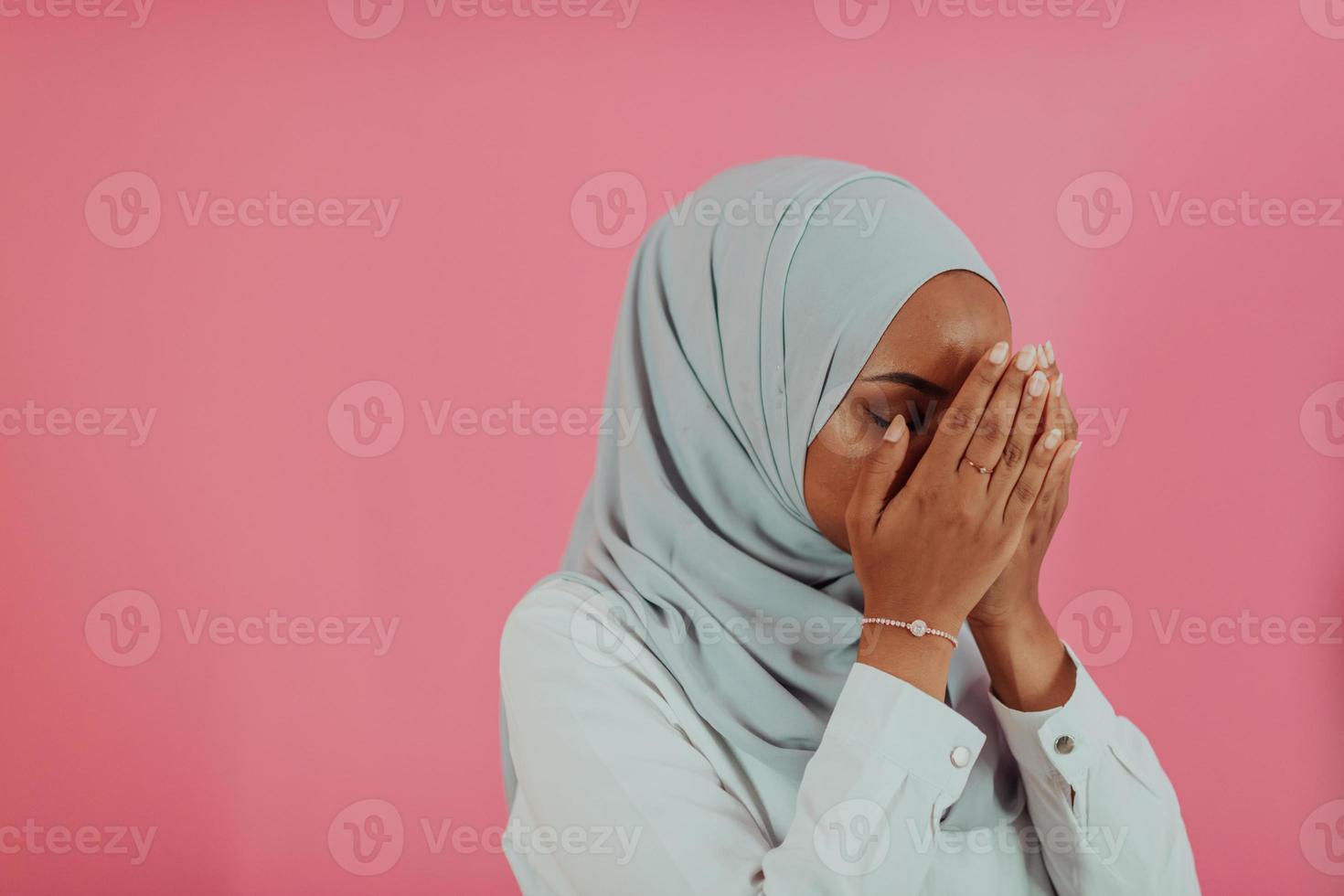 Modern African Muslim woman makes traditional prayer to God, keeps hands in praying gesture, wears traditional white clothes, has serious facial expression, isolated over plastic pink background photo