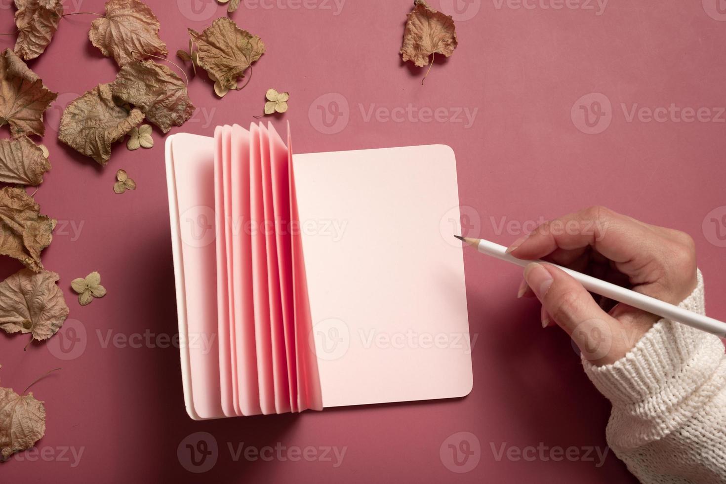 Office table desk with blank pink paper note pad and female hand wit pencil and fall leaves on colored background. Autumn flat lay composition with blank paper photo